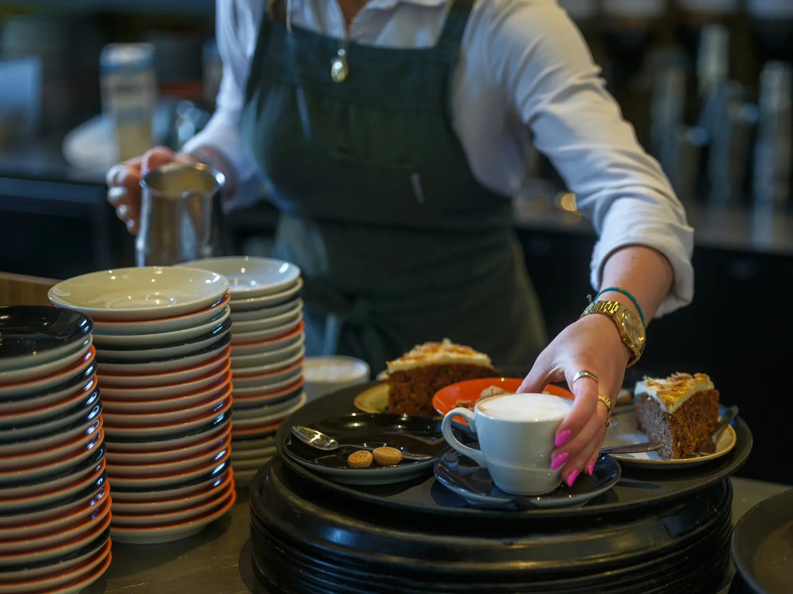 A server sets a cappuccino and slices of carrot cake on a tray at a busy cafe counter